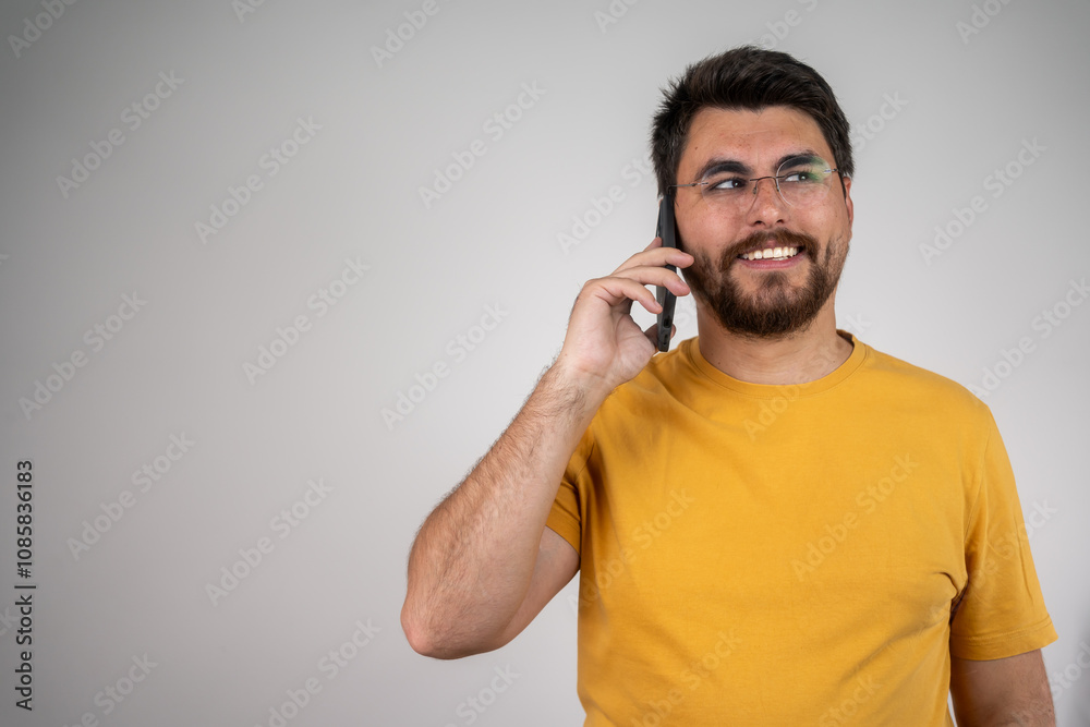 Smiling man with a beard, wearing a yellow t-shirt, is talking on the phone in front of an isolated background, looking relaxed and professional Smiling man with a beard, wearing a yellow t-shirt, is talking on the phone in front of an isolated background, looking relaxed and professional