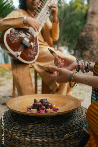 Indian musician in traditional clothes playing sitar festive season outdoor home. Woman in traditional saree hand lighting Diya lamp during Diwali festival. Happy greeting photo. Part of a series