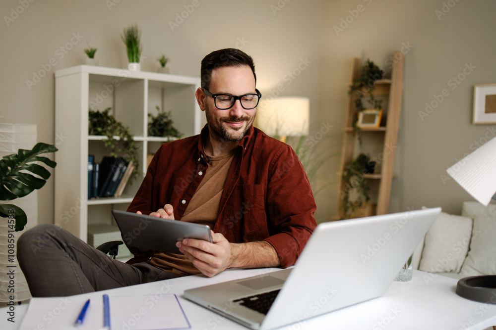 Happy businessman working remotely using laptop and tablet sitting at desk in home office Happy businessman working remotely using laptop and tablet sitting at desk in home office