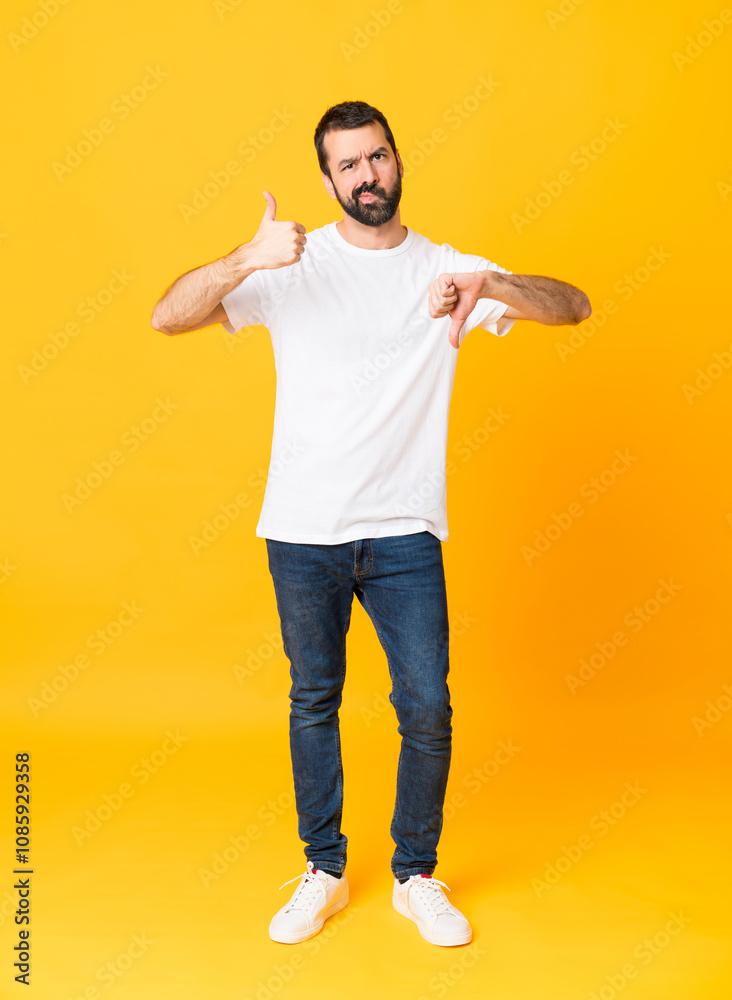 Full-length shot of man with beard over isolated yellow background making good-bad sign. Undecided between yes or not Full-length shot of man with beard over isolated yellow background making good-bad sign. Undecided between yes or not