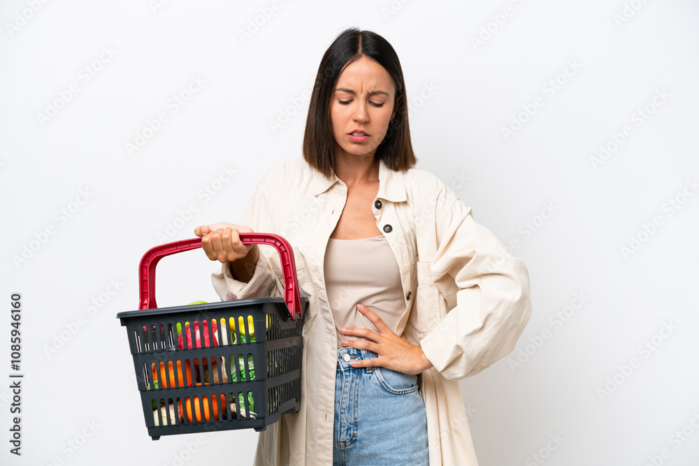 Young woman holding a shopping basket full of food isolated on white background suffering from backache for having made an effort Young woman holding a shopping basket full of food isolated on white background suffering from backache for having made an effort