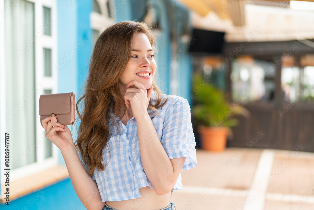 Young redhead woman holding a wallet at outdoors thinking an idea and looking side Young redhead woman holding a wallet at outdoors thinking an idea and looking side
