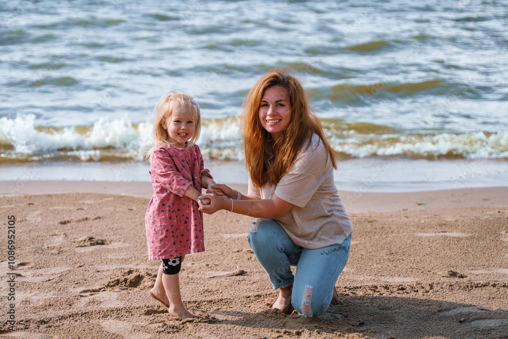 Little cute child toddler girl with a young mother on the beach on a cloudy day at sea. Little cute child toddler girl with a young mother on the beach on a cloudy day at sea.