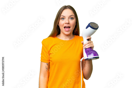 Young blonde woman holding a vertical steam iron over isolated chroma key background with surprise and shocked facial expression