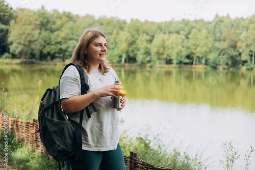 Happy smiling 30s women with backpack is holding thermos mug. Hot tea or other beverage on summer day. Lifestyle adventure concept. Woman drink cold water from the bottle photo