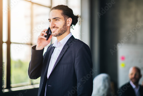 A young man dressed in a suit stands in an office, engaged in a phone conversation while smiling. Bright sunlight filters through large windows, creating a professional atmosphere.