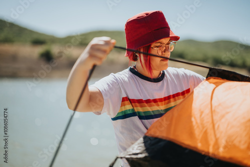 Young person assembling an orange tent by a lakeside on a sunny day. Ideal image for camping, adventure, and outdoor activities. photo