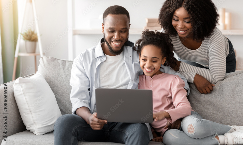 Family Weekend. Cheerful African American parents and their little daughter using laptop at home together, watching movie, photos or browsing internet, sitting on the comfortable couch in living room Family Weekend. Cheerful African American parents and their little daughter using laptop at home together, watching movie, photos or browsing internet, sitting on the comfortable couch in living room