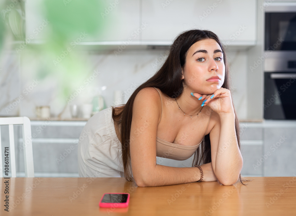 Bored young woman bending over table looking thoughtfully with smartphone placed next to her on kitchen-table Bored young woman bending over table looking thoughtfully with smartphone placed next to her on kitchen-table