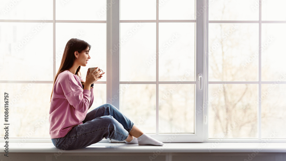 In The Morning. Profile of beautiful woman sitting on white windowsill at home, looking out of window and drinking hot coffee from cup. Happy lady taking break, holding mug with tea, copy space In The Morning. Profile of beautiful woman sitting on white windowsill at home, looking out of window and drinking hot coffee from cup. Happy lady taking break, holding mug with tea, copy space