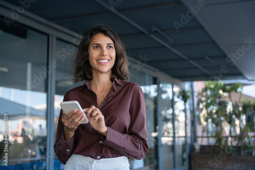 European business woman manager using cell phone mobile app standing outdoors at office. Latin Hispanic young female businesswoman freelancer working on smartphone, looking aside dreaming. Copy space photo