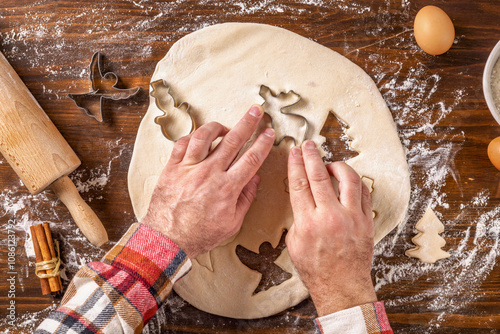 Top view of man's hands cutting out festive shapes from dough with cookie cutters on wooden table surrounded with baking ingredients. Cooking background