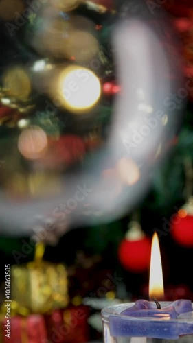 Close up of burning candle in focus with blurred decorated Christmas tree with blinking lights and blurred bulb with glow. Vertical video, details. Scene background.