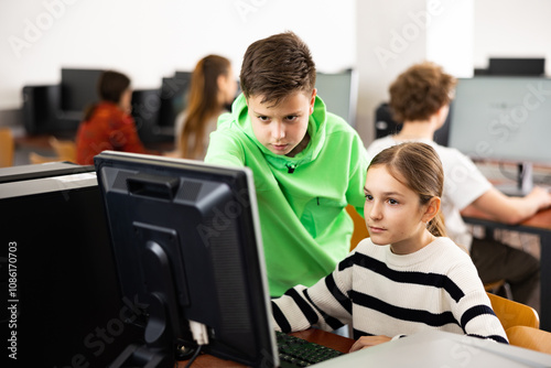 Smart interested preteen schoolboy helping cute girl classmate sitting at computer during informatics lesson..