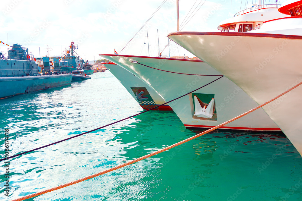 Bows of boats docked in a marina. The boats are tied with ropes to the dock, and the water is a clear turquoise color. In the background, there are more boats and some structures, harbor or port area