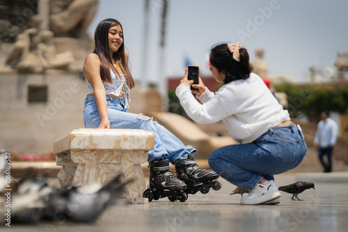Young woman taking photo of her skater friend sitting on bench in city square