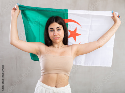 Happy young woman holding large flag of Algeria posing against background of light unicoloured wall photo