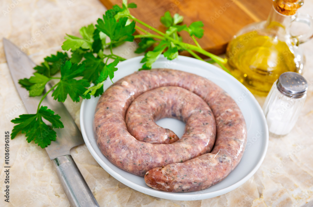 Fototapeta premium Coil of raw spiced butifarra sausage from minced pork prepared for frying lying on plate with knife fresh parsley, olive oil and salt. Traditional Spanish specialty