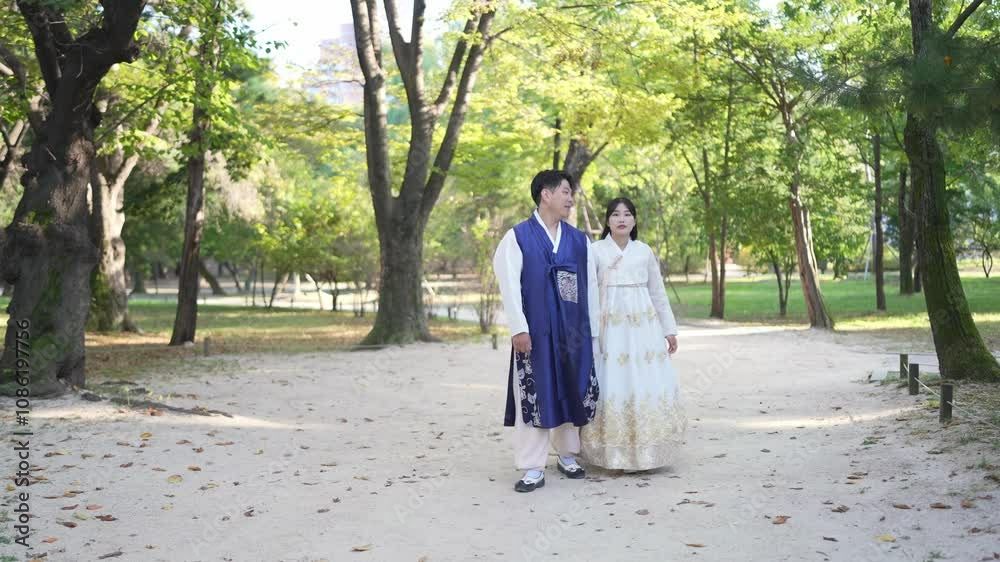 A Korean couple, a man in his 30s and a woman in her 20s, walk harmoniously through a park with historical buildings wearing Hanbok from Seoul, South Korea. A Korean couple, a man in his 30s and a woman in her 20s, walk harmoniously through a park with historical buildings wearing Hanbok from Seoul, South Korea.
