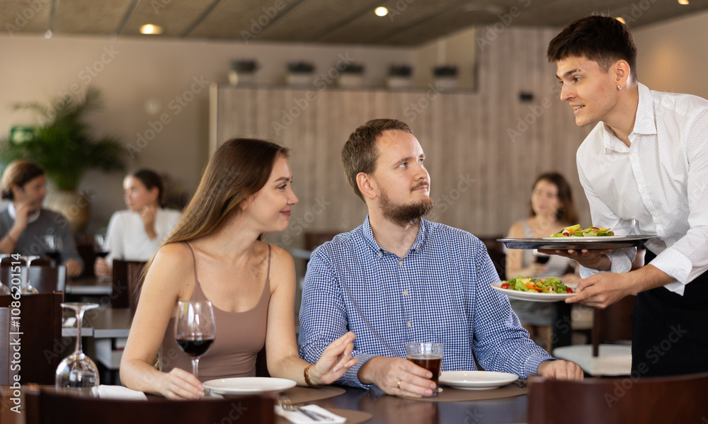 Friendly smiling waiter bringing dishes to guests of cozy restaurant Friendly smiling waiter bringing dishes to guests of cozy restaurant