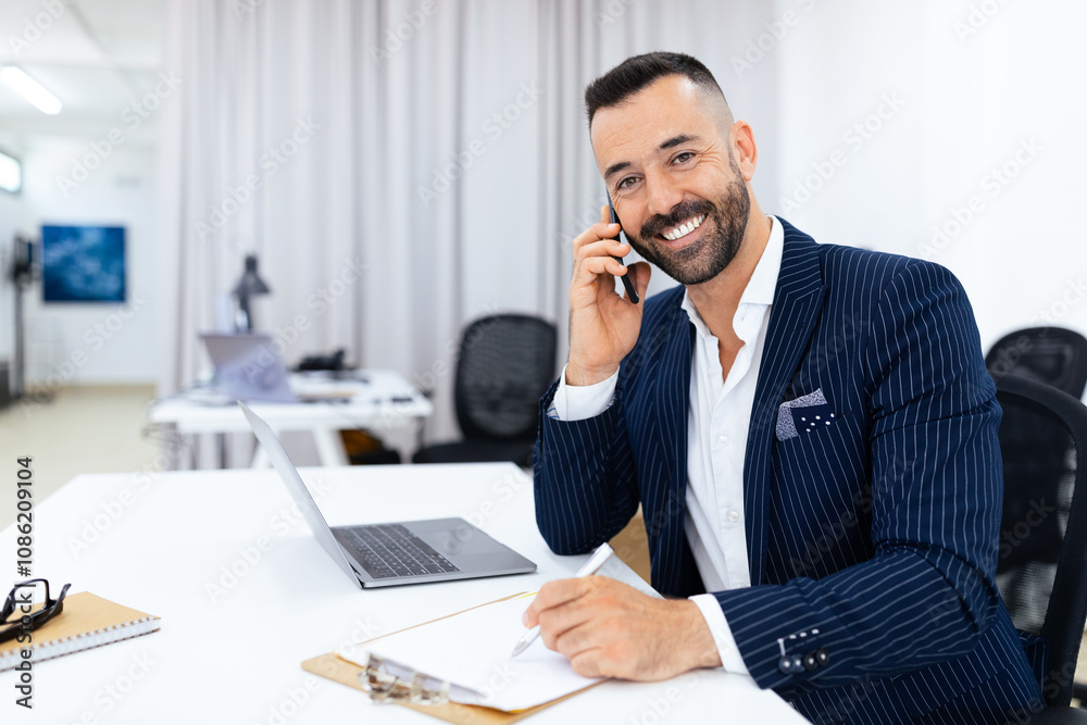 A man in a pinstriped suit smiles while talking on the phone, pen in hand, at a contemporary office desk. A laptop and notepad are nearby, indicating productivity in a professional environment. A man in a pinstriped suit smiles while talking on the phone, pen in hand, at a contemporary office desk. A laptop and notepad are nearby, indicating productivity in a professional environment.