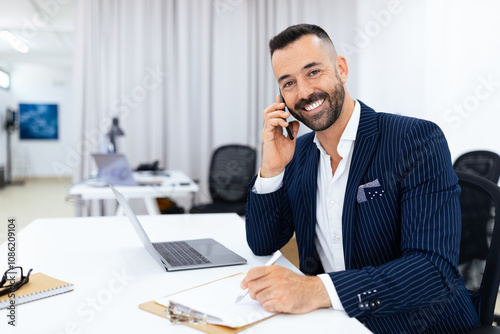 A man in a pinstriped suit smiles while talking on the phone, pen in hand, at a contemporary office desk. A laptop and notepad are nearby, indicating productivity in a professional environment.