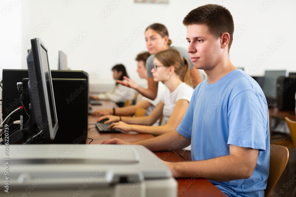 Side view on concentrated teenager male student sitting at library desk with pc, preparing for exam in computer class Side view on concentrated teenager male student sitting at library desk with pc, preparing for exam in computer class