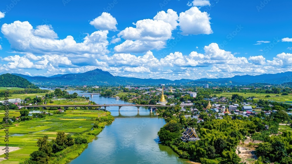 Fototapeta premium A vibrant landscape featuring a river, lush greenery, and mountains under a bright blue sky with fluffy clouds.