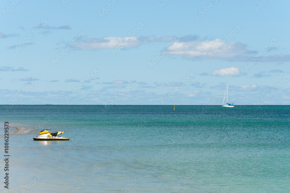 Obraz premium Near Perth, Western Australia: A peaceful coastal scene with a yellow and white jet ski floating in shallow turquoise waters. A sailboat is visible on the horizon beneath a partly cloudy sky.
