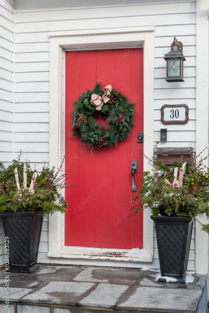 Naklejka premium The entrance to a white wooden house has a solid red door. A Christmas wreath hangs from the door. Two large black containers on either side are filled with fir, pine, and birch greenery.