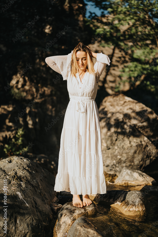 A woman in a long white dress stands on rocks by the riverside, embracing the calm and beauty of nature. A woman in a long white dress stands on rocks by the riverside, embracing the calm and beauty of nature.
