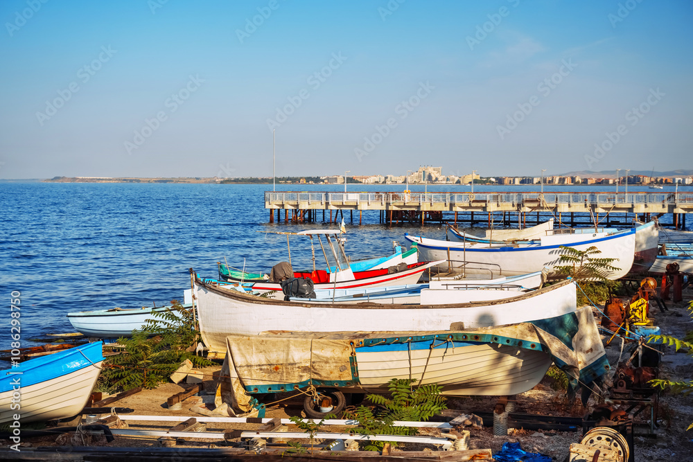 Brightly colored fishing boats are lined up along the shore, with a wooden pier extending into the serene sea. Brightly colored fishing boats are lined up along the shore, with a wooden pier extending into the serene sea.