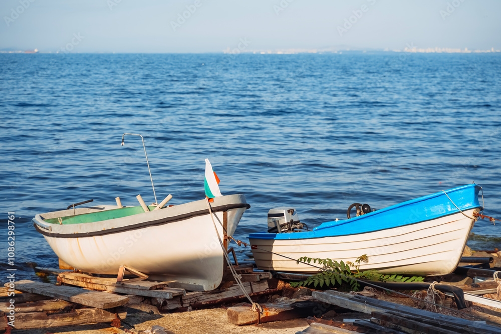 Naklejka premium Two small boats resting on wooden planks by the serene waters of a coastal location during daytime, with a gentle breeze ruffling the flag under a bright blue sky.