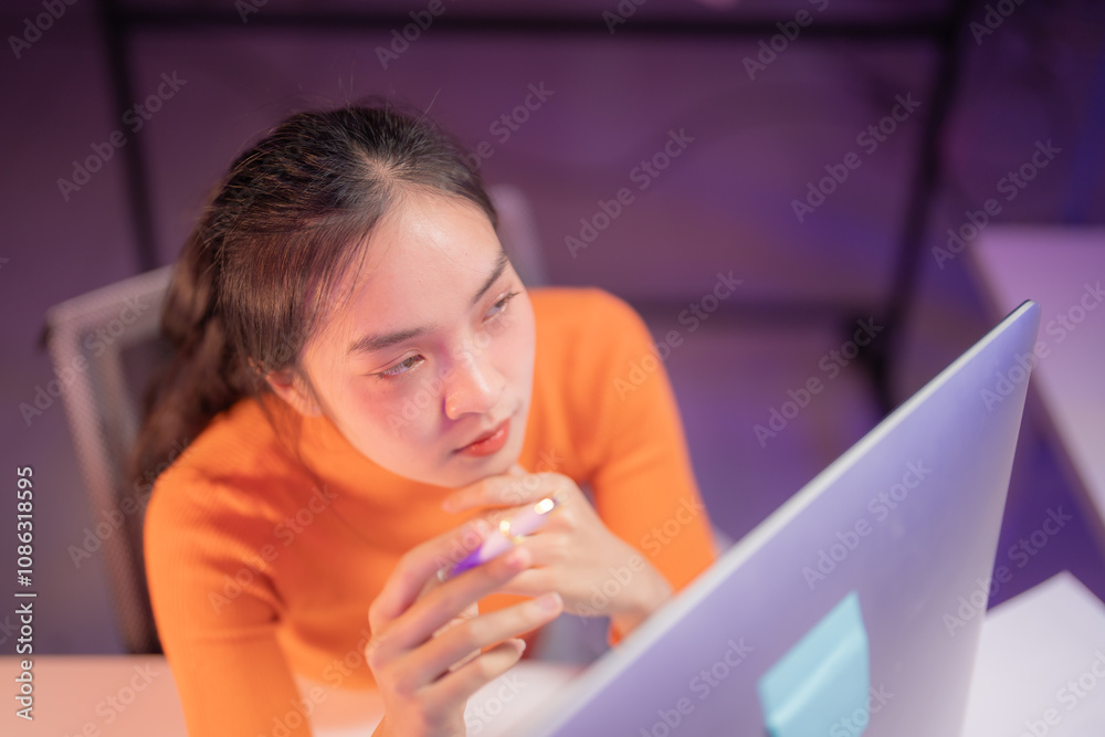 Young asian woman working remotely from home using her desktop computer, holding a pen and thinking about her next move in a dimly lit room with purple ambient light Young asian woman working remotely from home using her desktop computer, holding a pen and thinking about her next move in a dimly lit room with purple ambient light