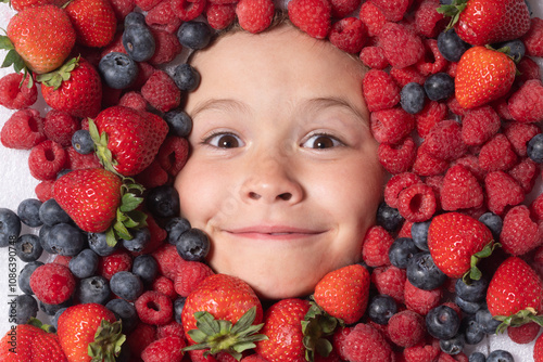 Funny fruits. Child face with berry frame, close up. Berries mix blueberry, raspberry, strawberry, blackberry. Assorted mix of strawberry, blueberry, raspberry, blackberry near child face. photo