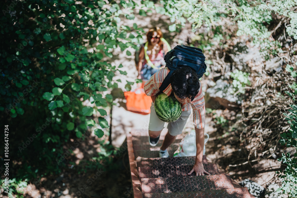 A man is climbing outdoor stairs carrying a watermelon, surrounded by green foliage, conveying a sense of adventure and the outdoors. A man is climbing outdoor stairs carrying a watermelon, surrounded by green foliage, conveying a sense of adventure and the outdoors.