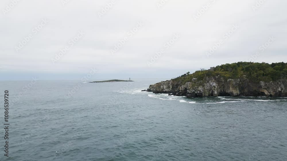 Rocky coastline with waves crashing near a small island and distant lighthouse on a cloudy day Rocky coastline with waves crashing near a small island and distant lighthouse on a cloudy day