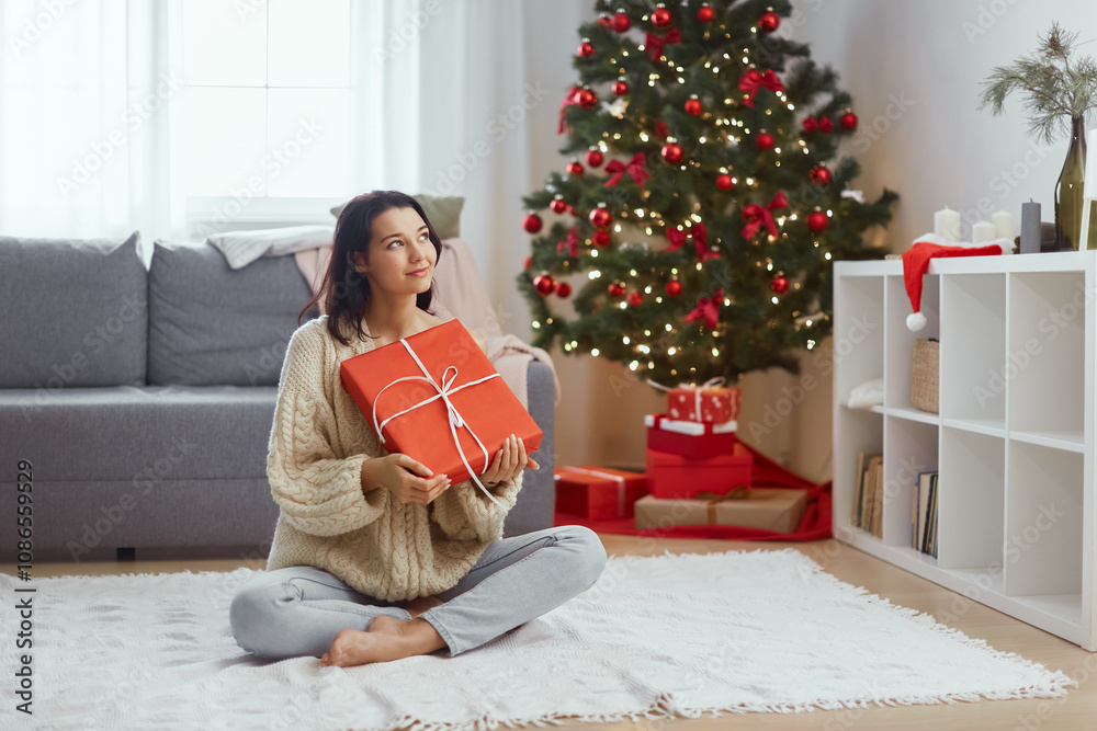 Young adult woman holding present in a wrapped red gift box, sitting in the living room at home in front of Christmas tree background. Thoughtful happy girl spending her winter holidays indoors Young adult woman holding present in a wrapped red gift box, sitting in the living room at home in front of Christmas tree background. Thoughtful happy girl spending her winter holidays indoors