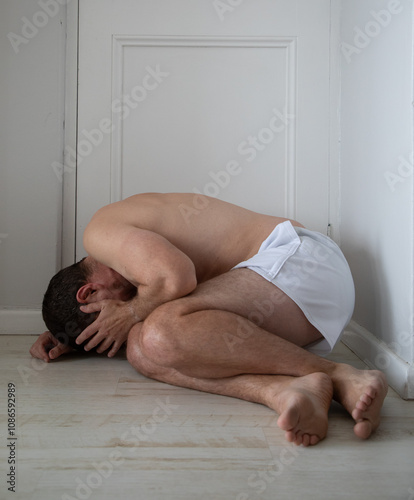 Caucasian male dressed in white shorts lying on the floor in an almost fetal position covering his face.  Concept for depression in middle aged men. 