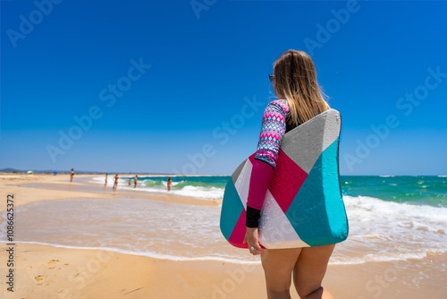 Beautiful young blonde woman in long sleeve swimsuit running with multicolored bodyboard on sandy beach by ocean shore on sunny day .Armona praia, Algarve, Portugal. Water sports in summertime. photo