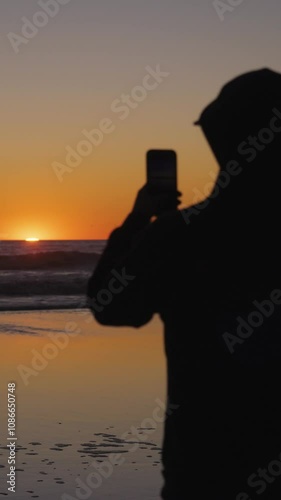 sillhouetted man films ocean sunset with mobil phone vertical video