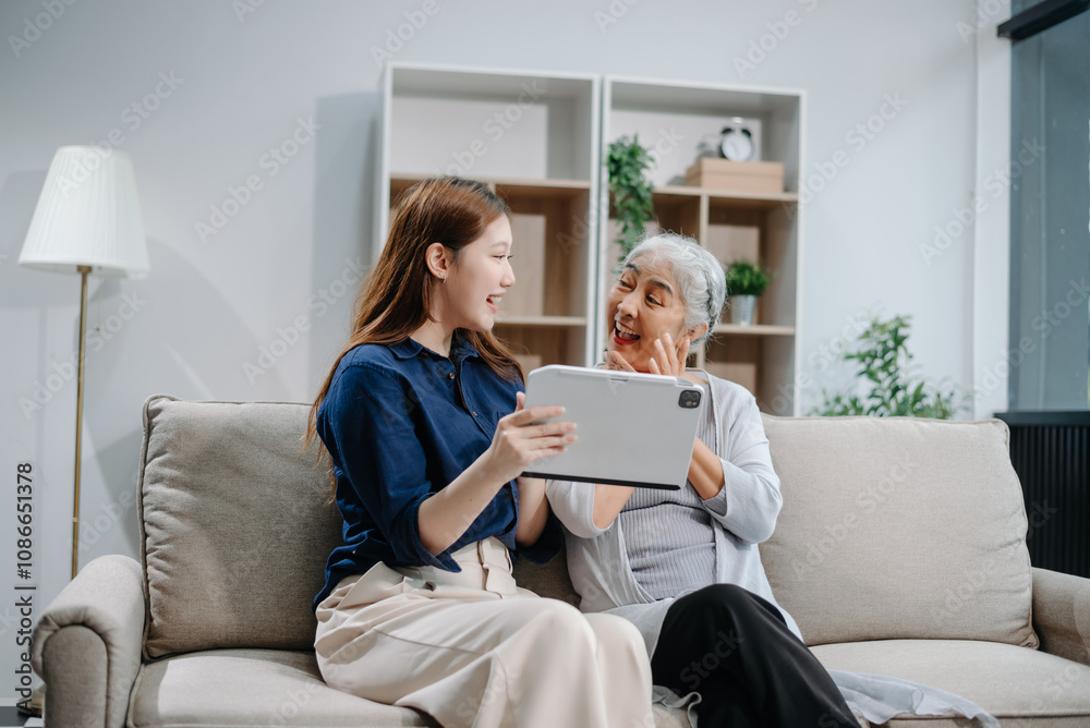 Happy adult Asian granddaughter and senior grandmother having fun enjoying talk sit on sofa in modern living room, using a laptop, tablet Happy adult Asian granddaughter and senior grandmother having fun enjoying talk sit on sofa in modern living room, using a laptop, tablet