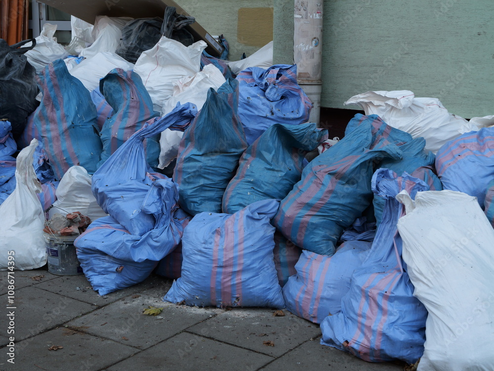 Piles of blue and white garbage bags stacked on a city sidewalk against a building wall, representing urban waste, cleanup, or construction debris removal Piles of blue and white garbage bags stacked on a city sidewalk against a building wall, representing urban waste, cleanup, or construction debris removal