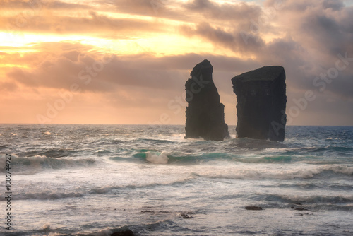 Twin rock formations rise dramatically from the ocean as the sun sets in the Azores Islands, creating a stunning seascape.