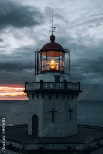 The lighthouse stands tall against a moody sky as the sun sets, casting a warm light over the ocean waves.