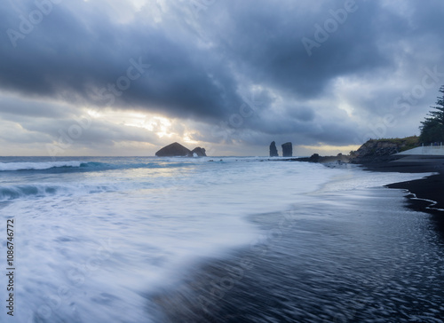 Waves crash softly on the black sand as the sun sets, illuminating the rocky coastline of the Azores Islands.
