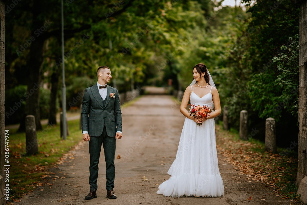 Bride and groom stand on a tree-lined path, gazing at each other with smiles, surrounded by greenery and autumn leaves, capturing a romantic wedding moment.. Bride and groom stand on a tree-lined path, gazing at each other with smiles, surrounded by greenery and autumn leaves, capturing a romantic wedding moment..