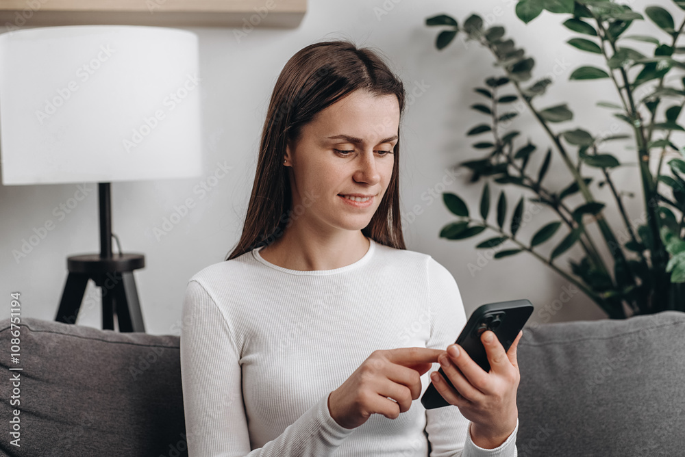 Cute young caucasian woman using smartphone sitting on comfortable sofa at home. Smiling beautiful girl using cell phone and texting. Technology, startup, new business and communication concept Cute young caucasian woman using smartphone sitting on comfortable sofa at home. Smiling beautiful girl using cell phone and texting. Technology, startup, new business and communication concept