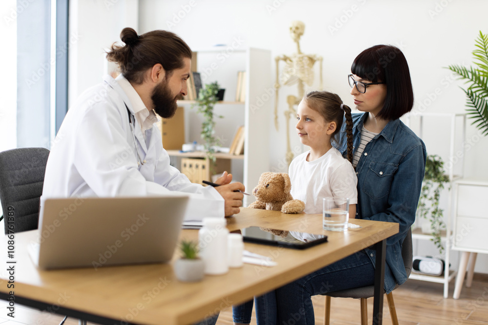 Caucasian male doctor interacts with young girl holding teddy bear during medical consultation in clinic. Mother, Caucasian woman, listens attentively beside daughter. Caucasian male doctor interacts with young girl holding teddy bear during medical consultation in clinic. Mother, Caucasian woman, listens attentively beside daughter.
