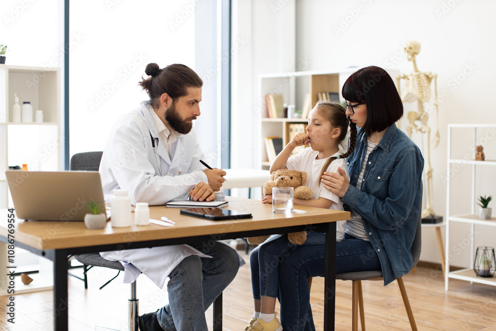 Caucasian male doctor examines Caucasian young girl coughing in clinic, writes medical prescription while Caucasian mother comforts girl. Caucasian male doctor examines Caucasian young girl coughing in clinic, writes medical prescription while Caucasian mother comforts girl.